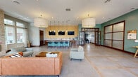 Located at 10531 Kings Row Dr in Avon, Indiana, this single-floor community clubhouse and lounge offers a generously sized, open-plan interior designed with contemporary elegance. The main gathering area is anchored by large-format tile flooring with inlaid accents, complemented by plush area rugs that define distinct seating zones furnished with leather sofas, modern accent chairs, and wood coffee tables. Oversized drum pendant lights hang from the elevated ceiling, casting a warm glow throughout the space.

A standout feature is the built-in bar, highlighted by a textured gold accent wall, pendant lighting, a marble-topped counter, and bar stools — ideal for social gatherings. Dual flat-screen TVs are mounted above the bar for added entertainment. On the opposite side of the room, a sleek linear fireplace set into a stacked-stone surround adds warmth and architectural interest.

Natural light pours in abundantly through multiple sets of French doors and transom windows along two walls, offering views of the surrounding greenery and easy access to outdoor areas. Teal accent walls and warm wood trim add a vibrant yet refined character throughout, making this a welcoming and functional communal space.