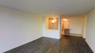 This single-floor space presents an open, airy layout designed for comfortable living. The main living area is finished with dark hardwood-style flooring that extends throughout, providing a warm, cohesive aesthetic against the clean white walls. A defining architectural feature is the partial dividing wall with a built-in pass-through window, which connects the living area to the kitchen zone while maintaining a sense of openness. The pass-through is accented with globe-style pendant lighting, adding a functional yet stylish touch.

The kitchen area, visible beyond the dividing wall, features modern flat-front cabinetry in a warm wood-toned finish and a light-colored countertop, offering a contemporary contrast to the darker flooring. Ceiling-mounted lighting throughout ensures the space remains well-lit. The open-plan configuration allows for flexible furniture arrangement in the living area, while the kitchen's layout appears efficient and practical. Overall, the space conveys a clean, modern aesthetic with thoughtful design details and a neutral palette suitable for a variety of interior styles.