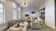 Located at 15155 Ollie Dr in Charlotte, NC, this single-story community clubhouse offers a thoughtfully designed gathering space that blends modern aesthetics with comfortable functionality. The main lounge is anchored by soaring ceilings that amplify the sense of openness, while an abundance of tall, grid-paned windows floods the interior with natural light and provides views of the surrounding neighborhood. The space features shiplap-accented walls in crisp white, contributing to a clean, contemporary atmosphere with subtle transitional charm. A striking geometric chandelier serves as a focal point overhead, complementing the curated selection of bold black-and-white abstract artwork displayed throughout. The lounge is furnished with multiple seating arrangements — channel-tufted sofas and armchairs in warm neutral tones paired with round marble-top and natural wood coffee tables — encouraging both social interaction and relaxed conversation. Patterned area rugs in earthy green and grey tones add texture and visual interest underfoot. Adjacent to the lounge, a dedicated business center provides granite-topped workstations, desktop computers, and printing equipment, all accented with elegant pendant lighting and coordinating abstract art. Glass-panel partitions and sliding doors seamlessly connect distinct zones within the space, maintaining an open and airy flow throughout.
