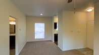 This single-story space presents a straightforward and functional layout with a clean, neutral aesthetic throughout. The main living area is finished with wall-to-wall carpet in a warm beige tone, providing a comfortable underfoot feel. Cream-colored walls and white baseboards give the space a bright, cohesive look, while a ceiling fan overhead ensures air circulation and comfort.

Natural light enters through a centrally positioned window, offering a view to the outdoors and keeping the room feeling open and airy. Recessed lighting supplements the natural light, ensuring the space remains well-lit throughout the day. A standard HVAC vent is visible in the ceiling, indicating a central climate control system.

An open passthrough connects the living area to the kitchen, which is visible in the background. The kitchen features dark cabinetry contrasting against lighter countertops and walls, with a dark appliance — likely a dishwasher — adding to the cohesive dark-toned kitchen design. The transition from carpet in the living area to hard flooring in the kitchen is clearly defined at the entryway, providing a practical delineation between the two spaces.