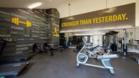 This fitness facility is thoughtfully designed to inspire and support a comprehensive workout experience across two distinct zones. The main gym area makes an immediate visual impact with bold, motivational wall graphics — including a striking "Stronger Than Yesterday" mural in yellow lettering — set against a dark, charcoal-toned backdrop. The space is outfitted with a wide range of cardio equipment, including stationary bikes, rowing machines, and ellipticals, as well as a dedicated strength training section featuring cable machines, a power rack, adjustable benches, and free weights. Durable rubber flooring, large wall-to-wall mirrors, and pendant lighting contribute to a focused, high-energy atmosphere. A separate multipurpose studio area offers a contrast in ambiance, featuring light wood-look flooring, soaring vaulted ceilings, and bright white walls that create an open, airy feel. A ballet barre with floor-to-ceiling mirrors lines one wall, making the space ideal for yoga, dance, or stretching sessions. Built-in cabinetry with a butcher block countertop adds functional storage, while large windows and a glass-paned door fill the room with natural light and provide views of an outdoor pool area.