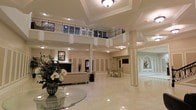 This impressive venue exudes sophistication and grandeur through its carefully crafted classical design. The centerpiece of the space is a dramatic double-height atrium, where a sweeping upper-level balcony with ornate wrought-iron railings overlooks the expansive main floor below. Polished marble flooring reflects the warm ambient lighting from recessed ceiling fixtures and elegant flush-mount lights, creating a luminous and inviting atmosphere throughout.

Stately columns punctuate the open floor plan, defining distinct areas while preserving the sense of openness and flow. Decorative wainscoting and paneled wall detailing add architectural refinement, complementing the coffered ceiling treatments above. A traditional fireplace anchors one end of the main gathering area, flanked by plush seating arrangements, while a separate dining area with upholstered chairs offers a more intimate setting.

An arched alcove with mirrored paneling at the far end of the space adds depth and visual interest, enhancing the overall sense of scale. Lush floral arrangements on a round glass accent table introduce a welcoming, decorative touch at the entrance. The overall ambiance is one of timeless elegance, making this space well-suited for formal gatherings, receptions, or upscale events.