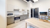 Located in Appleton, Wisconsin, this one-bedroom, one-bathroom unit offers a thoughtfully designed layout that maximizes every square foot. The space features a clean, modern aesthetic with crisp white walls, light-toned luxury vinyl plank flooring, and bold dark-framed doors and windows that add a striking contemporary contrast throughout.

The kitchen is a functional highlight of the unit, featuring two-tone cabinetry with white upper cabinets and gray lower cabinets, light countertops, a stainless steel refrigerator, a full range with microwave, and a single-basin sink. Tall ceilings with exposed dark beams lend an airy, loft-like character to the space, while a large window brings in natural light. A light wood dining table provides a convenient dining area just steps from the kitchen.

The bedroom is a cozy, well-appointed retreat with a wood-framed bed featuring under-bed storage drawers, neutral bedding, and botanical wall art that adds warmth to the white walls. A window offers natural light and an exterior view, completing the comfortable atmosphere.

The bathroom is accessible directly from the bedroom and features gray cabinetry, a marble-style countertop with an undermount sink, a large mirror, and light flooring that ties seamlessly into the rest of the unit's design palette.