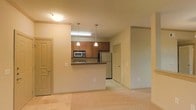 This single-floor space features an open-concept design that integrates the kitchen and living area into one cohesive layout. The kitchen is anchored by a raised peninsula counter with a dark countertop, which serves as a natural divider between the cooking and living spaces. Medium-toned wood cabinetry lines the kitchen walls, providing ample storage, while stainless steel appliances — including a refrigerator, microwave, and range — add a modern, functional touch. Two pendant lights hang above the peninsula, offering focused task lighting that complements the flush ceiling fixture in the living area. The flooring transitions from hard surface in the kitchen to wall-to-wall carpeting throughout the living space, contributing to a warm and comfortable ambiance. The neutral beige and tan color palette on the walls and floors keeps the interior feeling bright and versatile. A hallway visible beyond the kitchen leads to additional rooms, each accessed through interior doors consistent in style with the overall design. The layout is efficient and well-organized, making good use of the available space while maintaining an open, airy feel throughout.