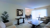 This single-floor residence offers a seamlessly connected living and dining area that creates a sense of openness and flow throughout the main living space. The living area features neutral-toned carpeting and is accented by a dark wood console table with ornate baluster legs, topped with decorative candle holders. A framed floral artwork in dark tones adds a sophisticated visual focal point to the wall. Adjacent to the living area, the dining space is distinguished by warm lighting from a classic chandelier that casts an inviting glow over a dark wood dining table and matching cross-back chairs. Large vertical blinds along the exterior wall allow abundant natural light to filter into the space, enhancing the airy, bright ambiance. The consistent use of soft, neutral wall tones and carpeting throughout creates a cohesive and calming aesthetic. Dark wood furnishings provide elegant contrast against the light-colored walls and floors, lending the space a balanced, classic character well-suited for both everyday living and entertaining.