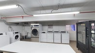 Located at 2525 Highland Avenue in National City, CA, this shared laundry facility offers a clean, functional space designed to serve its residents efficiently. The room is laid out on a single floor with a straightforward, utilitarian design, featuring white-painted walls and a smooth concrete ceiling with exposed utility conduits running overhead. Bright fluorescent lighting ensures the space is well-lit throughout.

The laundry area is equipped with a row of commercial-grade top-loading washers and dryers, alongside a large-capacity front-loading washer, providing ample options to accommodate varying laundry loads. A utility sink is conveniently situated at one end of the room, offering additional functionality for pre-treating garments or other cleaning tasks. A posted laundry room rules sign helps ensure shared use remains orderly and respectful for all users.

The flooring is a durable, easy-to-clean surface suited for the demands of a high-use communal space. A window on one wall allows for a degree of natural ventilation. The overall layout is practical and spacious enough to allow multiple users to move comfortably while attending to their laundry needs. This well-maintained facility reflects a commitment to providing essential, convenient amenities for residents of the property.
