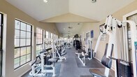 This fitness center is housed in a single-story facility with a striking vaulted ceiling that creates an open, airy atmosphere throughout the space. The cathedral-style roofline runs the full length of the room, adding a sense of height and volume that enhances the workout environment. Large, multi-pane windows line the walls on both sides, flooding the interior with natural light and offering views of the surrounding greenery and brick exterior buildings.

The flooring transitions between carpet and durable rubber matting in workout zones, providing appropriate surfaces for both machine-based and free-weight training. A ceiling fan is centrally mounted to support air circulation, complemented by recessed lighting for well-distributed illumination throughout.

The facility is comprehensively outfitted with a wide array of strength training machines, including cable systems, weight stack machines, and a multi-functional cable tower. A dedicated free weights area features a full rack of dumbbells positioned beneath large windows. Cardio equipment includes treadmills, a stair climber, and an elliptical machine, arranged along the perimeter to maximize floor space.

A wall-mounted television, a heart rate target chart, and a large mirror along one wall round out the amenities, making this a fully functional and well-designed fitness facility suited for a complete workout regimen. French doors at one end provide access to the exterior.