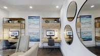 This single-floor space is thoughtfully designed to balance functionality and modern aesthetics. The workspace area serves as a focal point, anchored by a natural wood and metal open-frame desk unit that combines a work surface with overhead storage cabinetry. The warm, reclaimed-wood finish of the built-in desk system contrasts tastefully against the clean white walls, creating a visually grounded yet airy atmosphere. Recessed ceiling lighting provides practical illumination throughout, while a small task lamp on the desk adds warmth to the work area. Dark hardwood flooring runs throughout, adding depth and sophistication to the overall design. A pair of round mirrors with slim dark frames adorns one wall, enhancing the sense of space and reflecting light effectively. Abstract coastal-inspired artwork in shades of blue, teal, and white decorates the adjacent wall, introducing a calm, creative energy to the room. A neutral-toned accent chair nearby offers a comfortable seating option, rounding out the space's versatility. The overall ambiance is clean, contemporary, and well-curated, making it well-suited for both focused work and relaxed use.
