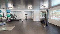 The Chace Lake Fitness Center in Hoover, Alabama, is a thoughtfully designed exercise facility offering a comprehensive range of workout equipment within a compact, efficiently utilized single-level layout. The interior features dark, high-gloss ceilings contrasted against crisp white walls, creating a sleek, modern aesthetic. Durable rubber flooring in a speckled dark tone covers the entire exercise area, providing both safety and functionality underfoot.

The space is divided into distinct workout zones. The cardio area houses multiple treadmills and an elliptical machine, positioned alongside a full-length mirrored wall that visually expands the room and assists users with form. The strength training section includes an Inspire cable machine, a multi-tiered dumbbell rack stocked with varied weights, kettlebells, a flat utility bench, a BOSU balance trainer, and exercise mats for floor work.

Geometric LED light fixtures illuminate the space alongside recessed ceiling lights, ensuring consistent brightness throughout. Multiple windows along the perimeter allow natural light to filter in, offering views of the surrounding outdoor pool area. A glass-paned door provides direct access to the pool, seamlessly connecting indoor fitness with outdoor amenities. Structural columns are neatly finished in white, integrating cleanly into the overall design.