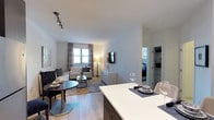 This one-bedroom unit in Quincy offers a thoughtfully designed open-concept floor plan that maximizes functionality and flow. The kitchen features a large white countertop island that serves as both a prep space and casual dining area, complemented by warm wood-toned cabinetry beneath. The island transitions naturally into a dedicated dining area, where a round dark-toned table provides an intimate setting for meals.

The living room extends toward a bright window dressed with elegant grey curtains, allowing natural light to fill the space. Light hardwood-style flooring runs throughout, lending warmth and visual continuity across the open layout. A mounted flat-screen television, a classic dresser, and tasteful décor complete the living area, which is anchored by a patterned area rug and a neutral sofa with coordinating accent pillows.

A corridor visible from the main living space leads to the bedroom, which offers a glimpse of a walk-in closet, adding practical storage. The unit maintains a clean, contemporary aesthetic throughout, with white walls, recessed lighting, and a track light fixture that enhance the bright, modern ambiance.