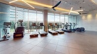 This fitness center is designed with both functionality and aesthetics in mind, offering a comprehensive workout environment across a single level. The cardio zone is a standout feature, lined with floor-to-ceiling windows that flood the space with natural light and provide expansive outdoor views of surrounding greenery and cityscape. Multiple treadmills, stationary bikes, a stair climber, and elliptical machines are arranged to take full advantage of these views, each equipped with individual display screens. Overhead, a striking geometric linear lighting installation with warm-toned LEDs is complemented by a large ceiling fan, ensuring a comfortable workout environment.

The strength training area is equally well-equipped, featuring a Smith machine with free weights, a cable machine, and a full rack of dumbbells arranged on tiered shelving. A wall of mirrors amplifies the sense of space and allows users to monitor their form. A dedicated amenity station adds a refined touch, incorporating a sleek dark tile accent wall, a marble-topped counter with built-in cabinetry, refrigerated beverages, and a mounted flat-screen television. Large-format tile flooring in a neutral tone ties the entire space together with a clean, modern finish.