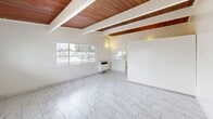 Located at 6565 Trigo Rd in Goleta, California, this compact yet thoughtfully designed single-floor unit offers a clean, airy aesthetic defined by standout architectural details. The most striking feature throughout the space is the rich, warm-toned wood plank ceiling accented by exposed beams, which adds character and a sense of height to the otherwise bright, white interior. Marble-look porcelain tile flooring runs consistently throughout, lending a cohesive and polished look underfoot.

The open main living area is bathed in natural light courtesy of multiple large windows dressed with horizontal blinds, offering views of the surrounding neighborhood greenery. The layout flows naturally into the kitchen, which is well-appointed with white cabinetry, light stone countertops, a double-basin sink, and stainless steel appliances including a gas range and full-size refrigerator. The wood ceiling continues into the kitchen, maintaining a warm, unified design language.

The bathroom is clean and functional, featuring marble-look tile flooring consistent with the rest of the unit, a full soaking tub with overhead shower, a single-sink vanity with white cabinetry, and a frosted window that allows natural light while ensuring privacy. The overall ambiance of the space is bright, minimalist, and inviting — well-suited for those seeking a low-maintenance lifestyle in a charming Goleta location.