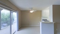 This single-floor space features an open-concept layout that connects the living area to the kitchen, creating a cohesive and functional floor plan. The living area is finished with wall-to-wall carpet in a neutral tone, complemented by warm beige walls and white baseboards that lend the space a clean, understated aesthetic. A ceiling fan with an integrated light fixture is centrally positioned overhead, providing both ventilation and ambient lighting. Natural light enters generously through a sliding glass door along one wall, which also provides access to an outdoor balcony, offering views of surrounding greenery. The kitchen is partially open to the living space, defined by a raised countertop bar that serves as a natural divider. White upper cabinetry lines the kitchen walls, and a gas range is visible, indicating a fully equipped cooking area. The textured ceiling adds a subtle detail consistent throughout the space. Overall, the layout is straightforward and practical, with a neutral color palette and functional finishes that create a bright, livable environment well-suited for everyday use.