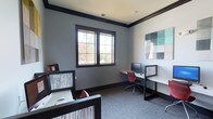 The Retreat offers a well-appointed workspace designed to support productivity in a comfortable, modern environment. The room features cool gray walls accented by dark crown molding and dark baseboards, creating a sleek, polished aesthetic. Carpeted flooring in a subtle pattern adds texture underfoot while contributing to a quiet atmosphere.

The space is equipped with multiple dedicated workstations, including built-in desk surfaces and freestanding computer desks, each paired with contemporary red chairs that add a pop of color against the neutral backdrop. Monitors are provided at the stations, making this a fully functional business center.

Natural light fills the room through a set of double windows with dark frames, offering views of the surrounding outdoor greenery. The windows are positioned to provide ample brightness without creating glare on the screens. A decorative teal and gray geometric artwork on the wall adds a modern artistic touch to the otherwise utilitarian space.

Divider panels with colorful accents help define individual workstation areas, offering a degree of privacy. Overall, the workspace balances functionality with contemporary design, making it an efficient and visually appealing environment.