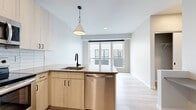 This contemporary single-floor unit showcases a clean, modern design with a neutral color palette and light hardwood flooring that flows seamlessly throughout the entire space. The open-concept kitchen serves as the heart of the unit, featuring light wood cabinetry with sleek black hardware, quartz countertops, and a full suite of stainless steel appliances including an electric range, over-the-range microwave, refrigerator, and dishwasher. A peninsula with an undermount sink and a stylish conical pendant light above adds both functionality and visual appeal. A crisp white subway tile backsplash completes the kitchen's polished look. The kitchen opens directly into the living area, which benefits from a large sliding glass door that invites natural light and provides access to an outdoor space. The unit includes multiple well-proportioned rooms, each finished with the same light hardwood flooring and neutral gray walls, offering a calm and versatile backdrop adaptable to a variety of uses. Ceiling fans are present in the rooms, enhancing comfort. The entry is anchored by a solid charcoal-toned door, adding a modern contrast to the bright, airy interior. Overall, the space presents a cohesive, move-in-ready aesthetic with contemporary finishes throughout.