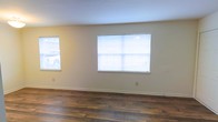 This single-story space presents a straightforward, functional layout with a clean and neutral aesthetic throughout. The main living area is finished with rich, dark hardwood-style flooring that adds warmth and visual contrast against the light-colored walls. The smooth, light beige walls provide a versatile backdrop suitable for a variety of décor styles.

Natural light is a notable feature of this space, with multiple windows fitted with white horizontal blinds allowing for adjustable light control. The larger window serves as a central focal point along one wall, drawing in ample daylight and contributing to an open, airy atmosphere. A secondary window on the adjacent wall further brightens the room.

The space maintains a minimalist character, with clean baseboards lining the perimeter and outlet placements conveniently positioned along the walls. The overall condition of the interior appears well-maintained, with fresh walls and flooring that suggest recent updates. The simple, uncluttered design makes it an adaptable space ready to accommodate a range of furnishing arrangements and personal touches.