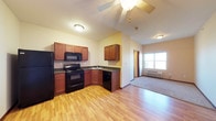This single-floor space showcases an open-concept layout connecting the kitchen and living areas seamlessly. The kitchen is equipped with rich medium-toned wood cabinetry, dark countertops, and black appliances including a refrigerator, electric range with overhead microwave, and dishwasher. An L-shaped counter configuration maximizes workspace and includes a sink positioned at the corner. Light hardwood-style flooring runs throughout the kitchen and main living area, adding warmth to the space. A standout architectural feature is the vaulted ceiling, which creates an enhanced sense of openness and volume. A ceiling fan is centrally mounted, providing both airflow and ambient lighting. The adjoining room, visible in the background, transitions to carpeted flooring and features a window that allows natural light to filter in, along with a wall-mounted heating unit beneath it. A doorway in the background hints at additional rooms beyond. The overall ambiance is functional and comfortable, with a neutral color palette complemented by warm wood tones throughout.