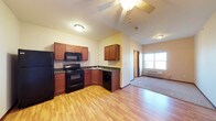 This single-floor space showcases an open-concept layout connecting the kitchen and living areas seamlessly. The kitchen is equipped with rich medium-toned wood cabinetry, dark countertops, and black appliances including a refrigerator, electric range with overhead microwave, and dishwasher. An L-shaped counter configuration maximizes workspace and includes a sink positioned at the corner. Light hardwood-style flooring runs throughout the kitchen and main living area, adding warmth to the space. A standout architectural feature is the vaulted ceiling, which creates an enhanced sense of openness and volume. A ceiling fan is centrally mounted, providing both airflow and ambient lighting. The adjoining room, visible in the background, transitions to carpeted flooring and features a window that allows natural light to filter in, along with a wall-mounted heating unit beneath it. A doorway in the background hints at additional rooms beyond. The overall ambiance is functional and comfortable, with a neutral color palette complemented by warm wood tones throughout.