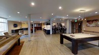 This single-floor communal lounge is a well-appointed recreational space designed to accommodate a variety of social activities. The open-plan layout is anchored by wood-look tile flooring that runs throughout, lending warmth and cohesion to the expansive interior. Recessed lighting is distributed evenly across the white ceiling, providing consistent illumination across all zones.

At the forefront of the space sits a full-sized pool table, serving as a central recreational focal point. Adjacent to it, a sleek kitchenette features dark cabinetry paired with a white countertop and sink, offering practical utility for gatherings. A high-top bar area, lined with bar stools, is set against a dramatic large-scale black-and-white mural depicting a group of musicians, creating a striking visual backdrop and adding a creative, artistic dimension to the space.

Toward the far end, a lounge area with casual seating and warm-toned walls offers a more relaxed setting. Structural columns are integrated throughout without disrupting the overall flow. The combination of entertainment amenities, open layout, and thoughtful design details makes this a versatile and inviting shared space.
