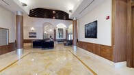 This sophisticated lobby presents a seamless blend of classical architecture and contemporary design. The expansive space is anchored by polished marble flooring with gold inlay accents, reflecting natural light that pours in through large glass entry doors flanked by tall, arched transoms. Lush potted fiddle-leaf fig trees frame the entrance, adding organic warmth to the refined setting.

Soaring ceilings feature an ornate latticed ceiling detail above a dramatic contemporary chandelier, lending grandeur to the central atrium. Structural columns are wrapped in warm walnut veneer, echoing the rich wood paneling that runs as a continuous wainscot throughout the space, complemented by matching wood-framed doorways and millwork details.

A welcoming seating area offers plush navy velvet sofas and armchairs arranged around a sleek round coffee table adorned with fresh floral arrangements. Curated contemporary artwork punctuates the white upper walls, adding visual interest. A staffed reception desk with an integrated wood surround provides a functional service point, while a built-in mailbox alcove reflects the space's practical, well-appointed design.