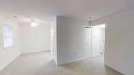 This single-floor space presents a bright, open layout characterized by its clean, neutral aesthetic. The interior is finished with wall-to-wall plush carpeting in a warm beige tone, lending a comfortable feel underfoot throughout the living areas. White walls and white ceilings create a sense of openness and maximize the effect of available light, giving the space an airy, welcoming ambiance.

A central dividing wall partially segments the interior, defining distinct functional zones while maintaining a connected, flowing layout. A ceiling fan is installed in the adjoining room, providing both air circulation and subtle overhead lighting. The space includes a closet with a full-height door, offering practical built-in storage. A thermostat is also visible on the wall, indicating individual climate control.

Doorways lead to additional areas, suggesting further rooms beyond the main living space. The overall design is straightforward and functional, with minimal architectural detailing. The consistent use of light finishes throughout contributes to a cohesive, understated interior that is well-suited for a variety of furnishing styles.