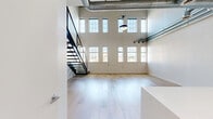 Located at 232 19th St NW in Atlanta's vibrant Atlantic Station neighborhood, this two-floor loft-style residence offers a striking blend of industrial character and modern finishes. The main level features an open-concept kitchen and living area anchored by light hardwood flooring throughout. The kitchen is outfitted with white cabinetry, quartz countertops, stainless steel appliances including a refrigerator, microwave, and range, and a large peninsula ideal for casual dining or entertaining. A half bathroom with a floating wood vanity and round mirror is conveniently located on the main floor. The living area is expansive, bathed in natural light from a dramatic bank of tall windows that frame views of the surrounding Atlanta cityscape. Soaring ceilings with exposed concrete beams, industrial ductwork, and conduit pipes lend the space a distinctive loft aesthetic. A sleek staircase with a black metal and cable railing leads to the upper level, which offers an open flexible space with the same hardwood flooring and continued industrial ceiling details. The upper floor also overlooks the main living area below, enhancing the sense of vertical scale. A full bathroom on the main level features marble-surround tub and shower, large-format tile flooring, and a marble-topped vanity. A utility room and closet complete the layout.