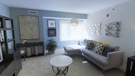 This single-floor space centers around a thoughtfully designed living room that combines contemporary style with a comfortable, welcoming ambiance. The room features a two-toned color scheme, with soft blue-gray accent walls contrasting against crisp white ceilings and lighter surrounding walls, creating a visually dynamic yet cohesive atmosphere. Plush wall-to-wall carpeting in a warm neutral tone runs throughout, adding to the overall sense of comfort.

The living area is anchored by a sectional sofa in light gray upholstery, accessorized with an assortment of decorative throw pillows in muted gold, charcoal, and patterned tones. A striped ottoman serves double duty as both a footrest and additional seating. A round white marble-topped coffee table with a sleek dark metal base sits at the center, providing both function and elegance.

Natural light filters generously through a large window fitted with vertical blinds, brightening the space. An arc floor lamp adds additional ambient lighting. Wall décor includes framed artwork and a textured metallic wall piece, while a large botanical canvas and a lush indoor plant introduce natural elements. A narrow console table along one wall completes the refined, curated aesthetic.