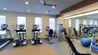 This fitness center is thoughtfully designed to provide a complete exercise environment within a single, open-plan space. The room is outfitted with a selection of cardio equipment, including elliptical trainers and an upright stationary bike, all positioned to take advantage of the natural light streaming in through multiple windows along the exterior wall. The bright, neutral-toned walls are accented by a bold, dark red upper border that adds a dynamic visual element to the space. A large wall-mounted mirror along one side of the room enhances the sense of openness while allowing users to monitor their form during workouts. Strength training equipment, including what appears to be a cable machine, is conveniently positioned near the mirror wall. The flooring features a combination of durable surfaces suited for both cardio and strength training activity. Recessed lighting throughout the ceiling, supplemented by a ceiling fan, ensures the space remains well-lit and well-ventilated. The layout is clean and uncluttered, maximizing usable floor space and allowing for comfortable movement between stations. Overall, the fitness center offers a practical and inviting environment for a wide range of exercise routines.