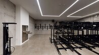 This single-level bike storage facility is designed with both functionality and modern industrial aesthetics in mind. The room is defined by its exposed concrete walls, which lend a raw, urban character to the space, contrasted by a polished light-toned floor that reflects the overhead lighting and enhances the sense of brightness and cleanliness.

Dominating the interior are multiple rows of heavy-duty, two-tier black steel bicycle racks capable of accommodating a substantial number of bikes. Additional wall-mounted bike hooks line one of the concrete walls, offering supplementary storage options for various bicycle types.

Overhead, a striking geometric arrangement of recessed linear LED strip lighting runs across the ceiling, providing even, efficient illumination throughout the room while adding a contemporary design element. A water fountain is conveniently located along one wall, serving the practical needs of cyclists.

Toward the rear of the space, a separate area with exposed concrete partitions and floor drains appears to function as a wash station or utility zone, providing a dedicated area for bike maintenance and cleaning. Access points are visible on multiple sides, ensuring straightforward entry and exit. Overall, the space balances industrial design with practical, high-capacity storage infrastructure.