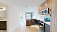 Located at 1923 Broadway Blvd in Kansas City, MO, this thoughtfully designed one-bedroom, one-bathroom unit offers a modern living experience across a single, efficiently laid-out floor. The kitchen is a standout feature, showcasing warm wood-grain cabinetry, bold teal-accented lower cabinets and appliances, a white quartz peninsula countertop, a marble-look tile backsplash, under-cabinet lighting, and light hardwood flooring. Stainless steel appliances, including a range and microwave, complete the contemporary cooking space. The bedroom is carpeted for comfort and features a large black-framed window that frames sweeping views of the Kansas City skyline, allowing abundant natural light to fill the room. Bi-fold doors open to a dedicated closet with built-in wire shelving, offering practical storage solutions. The bathroom is elegantly appointed with large-format marble-patterned floor tiles, a sleek dark wood vanity with gold hardware and a white countertop, a black-framed glass shower enclosure with teal tile detailing, and modern light fixtures above the mirror. The overall ambiance of the unit blends bold, contemporary design elements with functional, clean-lined spaces throughout.
