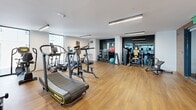Located at 1031 N Park Ave in Tucson, Arizona, this well-appointed fitness center offers a thoughtfully designed, single-floor workout facility. The space features warm hardwood-style flooring throughout, crisp white walls, and clean linear LED lighting that creates a bright, energizing atmosphere. Large floor-to-ceiling windows along multiple walls flood the interior with natural light and provide views of the surrounding cityscape, enhancing the open, airy feel of the room.

The facility is divided into distinct functional zones. The cardio section is equipped with a robust lineup of treadmills, ellipticals, stair climbers, stationary bikes, and a rowing machine — including equipment from brands such as TRUE and Invict. A dedicated strength and free weights area features a dumbbell rack, weight plates, cable machines, and a back extension bench, offering a comprehensive resistance training setup. Medicine balls and stability balls round out the functional fitness offerings.

A separate studio space is accessible through full-height glass doors and is lined with a textured wood slatted accent wall fitted with mirrors, making it ideal for stretching, yoga, or group fitness activities. The facility also provides convenient stairwell access, and the overall layout ensures ample open floor space for movement between equipment zones.