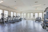 Located at 4251 Holly Springs Pkwy in Holly Springs, GA, this single-floor fitness center offers a spacious, thoughtfully designed workout facility. The open floor plan is anchored by distinctive herringbone-patterned flooring in a soft gray tone, lending the space a modern, polished aesthetic that complements its clean white walls and neutral trim.

Large windows line multiple walls throughout the facility, flooding the interior with abundant natural light and offering views of the surrounding community and outdoor pool area. Ceiling fans and linear pendant lighting provide additional comfort and illumination across the workout floor.

The cardio zone is well-appointed with multiple treadmills, elliptical trainers, a stationary bike, and a rowing machine — all positioned to take advantage of the natural light and outdoor views. The strength and functional training area features a multi-station cable machine, a dumbbell rack with a variety of weights, an adjustable bench, and a dedicated space for stretching and accessory work, complete with exercise balls and resistance tools stored on a wall-mounted rack.

Built-in glass-front cabinetry and a small beverage refrigerator provide added convenience. French doors and separate entry points offer easy access, while the overall layout ensures ample open space for movement and exercise variety. The bright, airy ambiance makes this a welcoming and motivating environment for all fitness levels.
