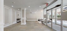 The lobby at 75 Clinton Pl in Hackensack, New Jersey presents a polished and welcoming environment designed with classic architectural details and practical amenities. Large-format natural stone tile flooring in muted gray and beige tones spans the entire space, lending it a grounded, sophisticated character throughout.

The walls are finished in warm neutral tones and accented with white wainscoting and decorative raised panel molding, creating a refined, traditional aesthetic. Crown molding runs along the ceiling perimeter, adding further architectural distinction. Recessed lighting provides even, ambient illumination throughout the lobby.

A comfortable seating area near the oversized glass storefront windows offers abundant natural light and views of the lush, tree-lined surroundings outside. The seating arrangement includes upholstered armchairs, a bench, and a glass-topped coffee table positioned over an ornate area rug, creating an inviting reception zone.

Adjacent to the entrance, a console table with decorative accessories and a round mirror serve as an elegant focal point. A dedicated mailbox area with built-in countertop shelving provides convenient resident services. A stainless steel elevator, framed by symmetrical panel molding, anchors one wall and ensures seamless accessibility. The overall ambiance is calm, elegant, and functional.