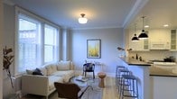 Located at 1520 Spruce Street in the heart of Philadelphia, PA, this single-floor residence offers a thoughtfully designed living space that blends comfort with contemporary style. The open-concept layout connects the living area seamlessly to the kitchen, creating an inviting and functional flow throughout the home. The living room is bathed in natural light from large windows dressed with blinds, illuminating the light hardwood flooring and soft, neutral-toned walls accented by classic crown molding. A flush-mount ceiling fixture adds a timeless touch overhead. The seating area is arranged around a glass coffee table and complemented by a mix of modern and accent chairs, giving the space a curated, welcoming feel. Adjacent to the living area, the kitchen features a breakfast bar with bar stools, pendant lighting, and dark countertops that contrast beautifully with white cabinetry — ideal for casual dining or entertaining. The cohesive design palette of whites, grays, and warm wood tones carries throughout, creating a bright and airy ambiance. Situated in Center City Philadelphia, this residence offers convenient access to the city's premier dining, cultural, and professional destinations.
