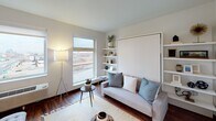 This thoughtfully designed single-floor unit maximizes space and functionality through a smart, open layout. The living area serves as the heart of the unit, featuring a built-in murphy bed flanked by open shelving, allowing the room to transition seamlessly between a comfortable lounge and a sleeping space. A large window floods the area with natural light and offers views of the surrounding landscape, complementing the warm hardwood flooring found throughout.

The kitchen is well-appointed with a two-tone design, pairing crisp white upper cabinetry with dark lower cabinets and dark granite countertops. Stainless steel appliances — including a refrigerator, electric range, and over-the-range microwave — provide a modern, cohesive aesthetic. Open shelving near the kitchen adds both display and storage utility.

A dedicated bathroom features a clean, minimalist vanity with a white countertop and dark cabinetry, maintaining the consistent design language of the unit. Conveniently situated nearby is an in-unit stacked washer and dryer, offering practical, day-to-day functionality without sacrificing space.

The overall ambiance is modern and efficient, with a neutral color palette, rich hardwood-style flooring, and carefully considered storage solutions that make this unit both visually appealing and highly livable.