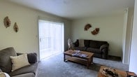 This single-floor space centers around a comfortably sized living room finished with wall-to-wall carpeting that adds warmth underfoot. A large sliding glass door dominates one wall, fitted with vertical blinds that allow for adjustable natural light, while also providing direct access to an outdoor area. The generous amount of light filtering through creates a bright, open atmosphere throughout the room. The living room accommodates ample seating and a central wooden coffee table, making it a functional and inviting area for relaxation or entertaining. The neutral-toned walls serve as a versatile backdrop, complemented by decorative metallic wall art that adds a subtle artistic touch. The textured ceiling adds a classic finishing detail to the space. Overall, the living room offers a practical and comfortable layout with an easy connection to the outdoors, making it well-suited for everyday living.