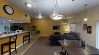 Located at 2524 Tamarack Ln in Indianapolis, IN, this single-floor space is thoughtfully designed to serve as a welcoming communal environment. The open layout connects several distinct functional zones, creating a cohesive and accessible flow throughout.

The main lounge area features warm golden-yellow walls, textured ceilings, and a mix of hard flooring and carpet. It is furnished with multiple seating arrangements, including leather sofas and upholstered chairs, centered around a wall-mounted flat-screen television. A decorative pendant light with a nature-inspired branch motif serves as a focal point overhead, complemented by additional flush-mount ceiling fixtures.

Adjacent to the lounge is a brighter sitting area distinguished by vaulted ceilings, pendant lighting, and a striking arched window paired with vertical blinds and sliding glass doors. This area allows for abundant natural light and views of the surrounding greenery outdoors.

The kitchen is a fully functional, commercially influenced space featuring stainless steel appliances — including a refrigerator, range, and microwave — set against warm wood cabinetry and exposed brick accent walls. A wide service counter with a laminate surface opens directly to the dining and lounge areas, facilitating easy interaction between the kitchen and gathering spaces.

Throughout the space, decorative touches such as wrought iron plant shelving, greenery, and layered lighting add warmth and character to this well-appointed community facility.