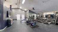 This single-level fitness center is designed to support a comprehensive workout experience within a spacious, open layout. The room features vaulted ceilings that rise to a notable height, lending an airy, open feel throughout the space. Flush-mounted fluorescent lighting and a ceiling fan ensure the area remains well-lit and ventilated during use.

The main workout area is lined with full-wall mirrors along one side, visually expanding the space while allowing users to monitor their form. The flooring throughout consists of durable rubber gym material, providing traction and impact absorption. Cardio equipment includes multiple treadmills, ICG spin bikes, and an elliptical or stair-climbing machine. Strength training is accommodated by a cable and weight machine setup positioned in a partially sectioned area with natural light from a window.

A separate changing area features grey carpeted flooring, a slatted bench, and a row of wall-mounted coat hooks for personal storage. The overall aesthetic is modern and functional, with grey-painted walls, white trim, and a clean, minimal design that maintains a professional fitness environment.
