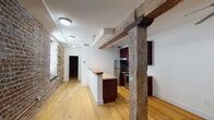 Located at 18 Horseford Rd in Lynchburg, Virginia, this single-floor space offers a compelling blend of historic architectural character and contemporary updates. The interior is immediately distinguished by its exposed original stone walls, which span entire sections of the living area and serve as a striking reminder of the building's storied past. Massive reclaimed timber posts and beams traverse the open layout, adding structural drama and rustic warmth throughout.

Light-toned hardwood flooring runs continuously across the main living areas, complementing the white walls and creating a bright, cohesive atmosphere. The kitchen features rich, dark cherry cabinetry paired with granite countertops, a peninsula with a breakfast bar, stainless steel appliances including a range, microwave, and refrigerator, and a double sink — offering a well-equipped cooking space with modern functionality.

A full bathroom is finished with clean white tile flooring and crisp white walls, maintaining a simple and practical aesthetic. In-unit laundry is conveniently accommodated with a stacked washer and dryer tucked neatly into a dedicated closet space. The stone-walled room at the far end of the unit presents a rare and evocative architectural feature, with floor-to-ceiling layered stonework evoking the building's original construction. Overall, the space delivers a memorable fusion of historic texture and thoughtful modern upgrades.