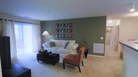 This single-floor residence offers a thoughtfully designed living space that balances comfort and style. The living room serves as the focal point of the home, anchored by a bold deep green accent wall that adds depth and character to the room. A plush white sofa paired with decorative throw pillows sits at the heart of the space, complemented by a tufted rust-colored armchair and a matching ottoman that introduce warm, earthy tones to the palette. A grid arrangement of decorative wall art adorns the accent wall, adding a curated, artistic touch. A classic floor lamp with a white shade provides soft ambient lighting, while sheer curtain panels allow natural light to filter gently into the room from an adjacent window. The neutral, light-toned carpeting runs throughout, creating a warm and inviting foundation that ties the furnishings together seamlessly. A wall-mounted HVAC vent and a small side table with accent décor round out the functional and aesthetic details of the living area. The overall ambiance is welcoming and relaxed, with a cohesive color scheme that blends cool greens, warm neutrals, and rich earth tones for a comfortable, homey atmosphere.