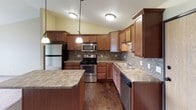 This single-floor home offers a thoughtfully designed layout with warm, neutral tones and quality finishes throughout. The kitchen is a standout feature, showcasing rich medium-toned wood cabinetry, granite countertops, and a mosaic tile backsplash that adds texture and visual interest. Stainless steel appliances — including a refrigerator, gas range, microwave, and dishwasher — provide a sleek, modern touch. Pendant lighting over the kitchen island and flush-mount ceiling fixtures illuminate the space, while dark hardwood flooring grounds the room with warmth. The kitchen opens seamlessly toward a carpeted living area, creating an inviting, connected flow between spaces. The bedroom is a calm and comfortable retreat, featuring plush wall-to-wall carpeting, soft neutral walls, and a window fitted with blinds that allows natural light while maintaining privacy. The full bathroom mirrors the kitchen's design sensibility, featuring granite countertops on rich wood vanity cabinetry, a full tub and shower combo, tiled flooring in a stone-like pattern, and dark-framed mirrors and fixtures for a cohesive, polished look. Throughout the home, consistent use of warm wood tones, neutral wall colors, and quality materials creates a unified and welcoming ambiance.