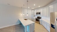 This single-story residence at Herrons Ferry Road in Rock Hill, South Carolina, offers a well-designed layout with contemporary finishes throughout. The kitchen serves as a central highlight, featuring light gray shaker-style cabinetry extending to the ceiling, white quartz countertops, and a large central island with an integrated sink and seating overhang. Three pendant lights hang above the island, complementing recessed ceiling lighting. Stainless steel appliances, including a range, over-the-range microwave, and French door refrigerator, complete the space. Light wood-look flooring runs through the kitchen and flows into the adjacent living area, which is brightened by a large window and glass-paned exterior door offering outdoor views. A ceiling fan provides additional comfort. The bedroom areas feature plush carpeting, ceiling fans, and generous natural light from multiple windows framed by white blinds. The primary bedroom is complemented by a spacious walk-in closet outfitted with multi-level wire shelving and hanging rods. The bathroom includes light wood-tone tile flooring, a single-sink vanity with white countertop, a large mirror with bar lighting, and grab bars for added accessibility.