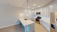 This single-story residence at Herrons Ferry Road in Rock Hill, South Carolina, offers a well-designed layout with contemporary finishes throughout. The kitchen serves as a central highlight, featuring light gray shaker-style cabinetry extending to the ceiling, white quartz countertops, and a large central island with an integrated sink and seating overhang. Three pendant lights hang above the island, complementing recessed ceiling lighting. Stainless steel appliances, including a range, over-the-range microwave, and French door refrigerator, complete the space. Light wood-look flooring runs through the kitchen and flows into the adjacent living area, which is brightened by a large window and glass-paned exterior door offering outdoor views. A ceiling fan provides additional comfort. The bedroom areas feature plush carpeting, ceiling fans, and generous natural light from multiple windows framed by white blinds. The primary bedroom is complemented by a spacious walk-in closet outfitted with multi-level wire shelving and hanging rods. The bathroom includes light wood-tone tile flooring, a single-sink vanity with white countertop, a large mirror with bar lighting, and grab bars for added accessibility.