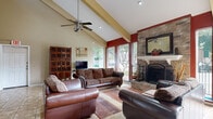 This single-floor space offers a well-appointed and inviting open layout designed for comfort and social gathering. The most commanding architectural feature is the soaring vaulted ceiling, accentuated by exposed diagonal beams and a ceiling fan that draws the eye upward, creating a strong sense of openness and volume. Bold accent walls in deep red contrast with neutral beige tones throughout, lending the space a warm yet dynamic character.

At the heart of the room stands a full-height stone fireplace, crafted from stacked natural stone and topped with a white mantel. A decorative mirror above the mantel further enhances the fireplace's dramatic presence. Arranged around the fireplace is a seating area with leather and upholstered sofas atop a richly colored area rug laid over neutral tile flooring, which extends throughout the space.

Large floor-to-ceiling windows and glass doors along one wall flood the interior with natural light and provide views of the surrounding outdoor greenery. The entryway features a white front door with an oval glass insert, tile flooring, and decorative wall art, creating a welcoming first impression. Additional interior doors and HVAC vents reflect thoughtful utility throughout. A television and display cabinet complete the functional living area, making this space equally suited for relaxation and social engagement.