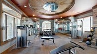 Located at 1000 Avonlea Pl in Woodstock, GA, this fitness center is a thoughtfully designed exercise facility that combines functional layout with upscale architectural details. The space is anchored by a dramatic coffered ceiling medallion with a wood-paneled inset and a ceiling fan, all set against a rich terracotta-colored ceiling that lends warmth and character throughout.

The facility is organized into distinct workout zones. A cardio area lines one side of the room with a row of treadmills and elliptical machines, positioned beneath large windows that invite natural light and outdoor views. Adjacent to this, a free weights zone features a full dumbbell rack, medicine ball storage, and adjustable benches, accented by a rustic wood-paneled wall that adds texture and visual interest. Floor-to-ceiling mirrors expand the sense of space and allow users to monitor their form.

The strength training area offers cable and selectorized machines, while open floor space with rubber matting provides room for stretching and functional exercises. The flooring transitions between large-format tile and carpet tile, defining each zone clearly. Elegant glass-paned double doors serve as the main entry, and a red accent chair near the entrance adds a pop of color. Recessed lighting throughout ensures a bright, well-lit environment suited for all-hours use.