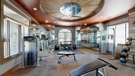 Located at 1000 Avonlea Pl in Woodstock, GA, this fitness center is a thoughtfully designed exercise facility that combines functional layout with upscale architectural details. The space is anchored by a dramatic coffered ceiling medallion with a wood-paneled inset and a ceiling fan, all set against a rich terracotta-colored ceiling that lends warmth and character throughout.

The facility is organized into distinct workout zones. A cardio area lines one side of the room with a row of treadmills and elliptical machines, positioned beneath large windows that invite natural light and outdoor views. Adjacent to this, a free weights zone features a full dumbbell rack, medicine ball storage, and adjustable benches, accented by a rustic wood-paneled wall that adds texture and visual interest. Floor-to-ceiling mirrors expand the sense of space and allow users to monitor their form.

The strength training area offers cable and selectorized machines, while open floor space with rubber matting provides room for stretching and functional exercises. The flooring transitions between large-format tile and carpet tile, defining each zone clearly. Elegant glass-paned double doors serve as the main entry, and a red accent chair near the entrance adds a pop of color. Recessed lighting throughout ensures a bright, well-lit environment suited for all-hours use.