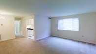 This single-floor space features an open and functional layout connecting a spacious living area to a dedicated kitchen. The living area is finished with wall-to-wall beige carpeting, white walls, and clean white baseboards, creating a bright and neutral atmosphere. A window fitted with horizontal blinds allows natural light to filter into the room, enhancing its airy feel. Overhead, a ceiling-mounted HVAC vent provides climate control throughout the space.

Adjacent to the living area, the kitchen is visible through an open passageway, offering a seamless connection between the two spaces. The kitchen features white cabinetry, a white gas range, and tile or vinyl flooring, presenting a clean and practical cooking environment. The cabinetry provides ample storage, while the layout allows for efficient use of the available space.

Overall, the unit maintains a simple and well-maintained aesthetic with a consistent color palette of whites and neutral tones throughout. The open sightline between the living area and kitchen contributes to a connected and cohesive feel across the entire floor plan.
