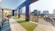 Located at 90 102nd Ave NE in the heart of Bellevue, Washington, this rooftop amenity space offers a sophisticated outdoor experience defined by modern design and premium finishes. The single-level space is clad in sleek, polished stainless steel panels that form a striking architectural core housing an elevator lobby, providing seamless vertical access to the rooftop.

The outdoor deck is thoughtfully designed with a putting green featuring multiple hitting stations, framed by bold dark metal columns that create a covered practice area with an open, airy feel. The turf stations face outward toward panoramic views of the Bellevue skyline, with surrounding high-rise developments forming an impressive urban backdrop. Decorative river rock beds border structural elements, adding a natural textural contrast to the polished metal and concrete palette.

Ample lounge seating is arranged throughout the deck, inviting occupants to relax and take in the expansive views of the city and surrounding tree canopy. Safety railings line the perimeter, and designated emergency exit access points are clearly marked. The overall ambiance is clean, contemporary, and distinctly urban, making this rooftop an exceptional amenity space suited for leisure and recreation.