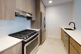 Located at 1277 E 14th St in the Flatbush neighborhood of Brooklyn, NY, this modern single-floor residence showcases clean architectural lines and high-quality contemporary finishes throughout. Light-toned hardwood floors run through the living areas and bedrooms, creating a warm and cohesive aesthetic that complements the crisp white walls.

The open-concept living and dining area is bathed in natural light, with large windows offering views of the surrounding neighborhood. A set of glass doors leads to a private balcony, further enhancing the sense of openness. An adjoining bedroom features its own oversized window with an urban backdrop and a wall-mounted radiator.

The kitchen is a standout feature, designed with wood-toned cabinetry, a patterned geometric tile backsplash, and a suite of stainless steel appliances including a gas range and full-size refrigerator. A peninsula with a contrasting weathered-wood finish and a matte black sink provides additional prep space and a natural transition into the living area, with light tile flooring completing the culinary space.

The bathroom is thoughtfully appointed with charcoal penny-round mosaic floor tiles, large-format neutral wall tiles, a wood-grain vanity with an integrated white countertop, and a recessed medicine cabinet mirror, delivering a refined and modern feel.