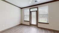 This single-floor residence offers a thoughtfully designed layout with a cohesive contemporary aesthetic throughout. The entryway opens directly adjacent to the kitchen, which features warm-toned shaker-style cabinetry, granite countertops, a subway tile backsplash, stainless steel appliances including a refrigerator, electric range, and built-in microwave, and a pendant light-adorned breakfast bar that connects seamlessly to the living area.

The living space is bright and open, benefiting from multiple windows flanking a private exterior door that provides views of surrounding greenery. Crown molding, a ceiling fan, and light wood-look luxury vinyl flooring contribute to a polished, cohesive feel. A dedicated dining or flex area mirrors this aesthetic, offering additional natural light.

The bedrooms are carpeted for comfort and include ceiling fans and ample natural light from window treatments. At least one bedroom features access to a walk-in closet equipped with built-in shelving and hanging racks. The bathroom is finished with granite countertops, warm wood cabinetry with brushed nickel hardware, a large framed mirror, and a full tub-shower combination with clean white surround.